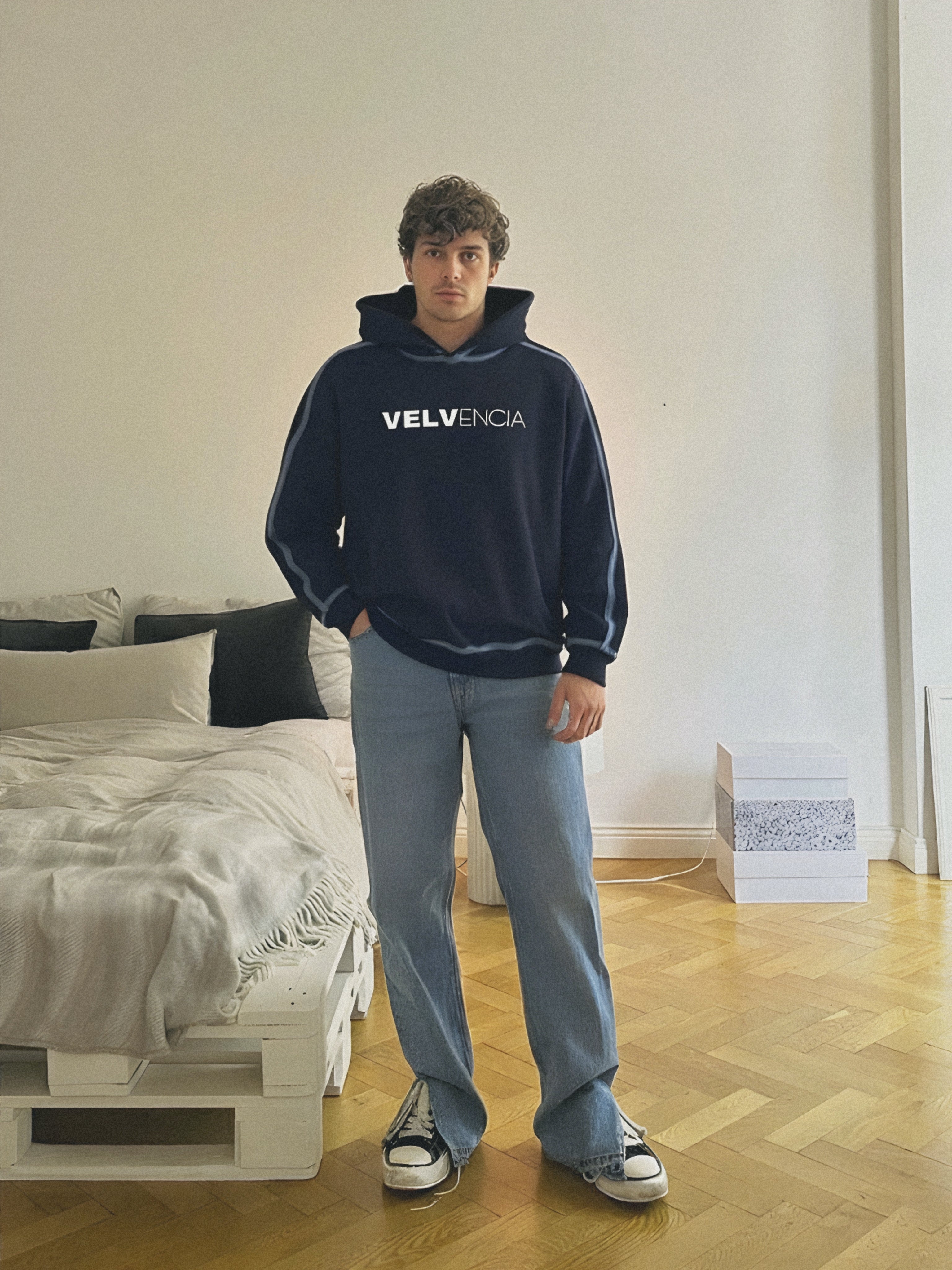 Person wearing a navy hoodie with 'VELVENIA' logo standing in a room with a bed and wooden floor.