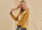 Woman wearing a yellow and gray striped sweater with a graphic design, blue jeans, and beige sneakers against a beige wall.