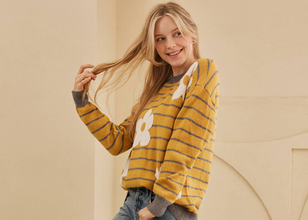Woman wearing a yellow and gray striped sweater with a graphic design, blue jeans, and beige sneakers against a beige wall.